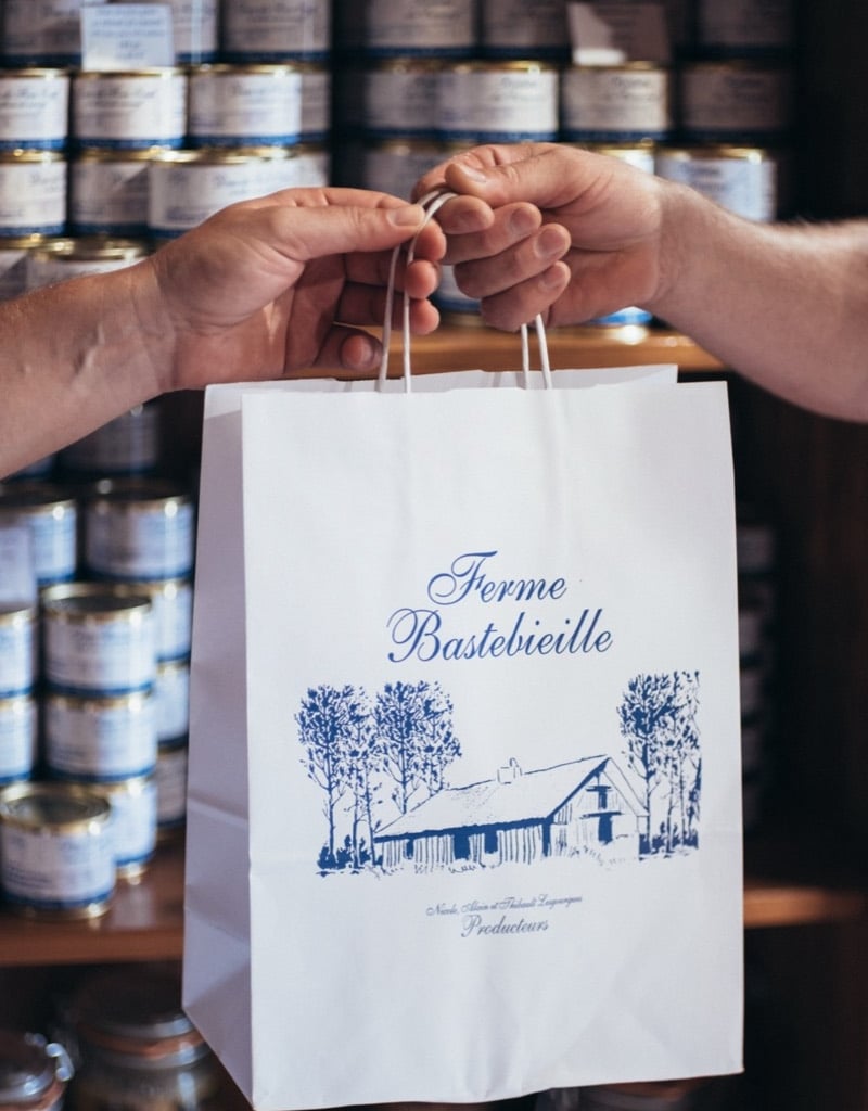 sac cadeau boutique ferme bastebieille Deux mains tendent un sac cadeau de la Ferme Bastebieille devant les étagères de la boutique